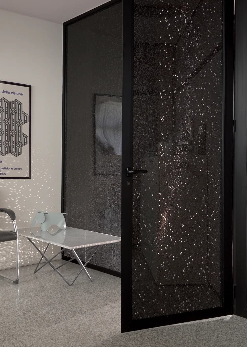 Koros door in a black aluminium frame with a dark bubble patterned panel beside a framed poster and a table